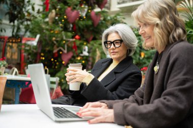 İki son sınıf öğrencisi dizüstü bilgisayarda çalışıyor ve dışarıda bir Noel ağacının yanında kahve içiyor.