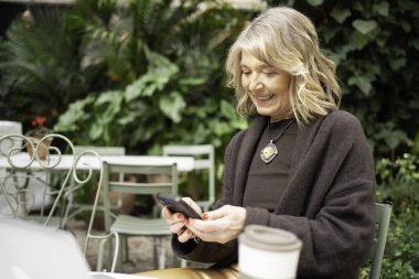 Olgun bir kadın mutlu bir şekilde dışarıda telefon kullanıyor, bitkilerle dolu bir ortamda teknolojinin tadını çıkarıyor.