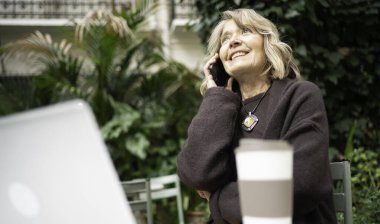 Bir telefon görüşmesi yaparken gülümseyen bir kadın, açık bir kafede oturuyor.