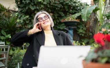 Dışarıda dizüstü bilgisayar üzerinde çalışırken akıllı telefonla iletişim kuran olgun bir kadın.