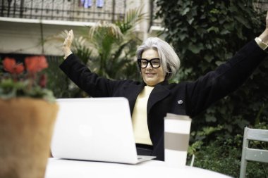 Üst düzey kadın dışarıda dizüstü bilgisayarla çalışırken kollarını kaldırarak online başarıyı kutluyor.