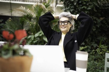 Gri saçlı kadın gülümsüyor, kollarını açıyor, laptoplu ve kahveli kahve keyfi yapıyor.