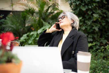 Gri saçlı, akıllı telefonlu, laptoplu ve kafede kahve içen olgun bir kadın.