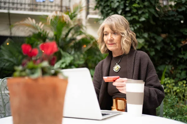 Yaşlı bir kadın dışarıda otururken kredi kartı ve laptopuyla online alışveriş yapıyor.