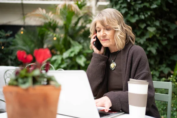 Üst düzey bir kadın akıllı telefondan iletişim kuruyor ve açık bir kafede dizüstü bilgisayarla çalışıyor.