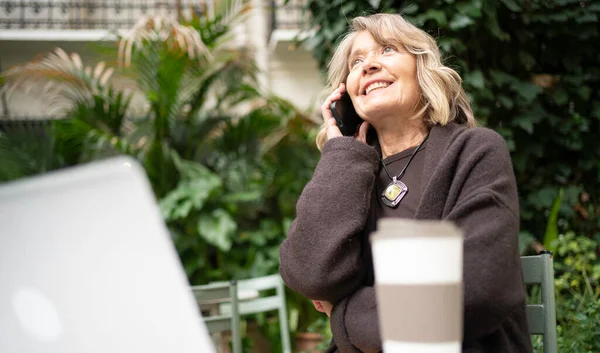 Bir telefon görüşmesi yaparken gülümseyen bir kadın, açık bir kafede oturuyor.