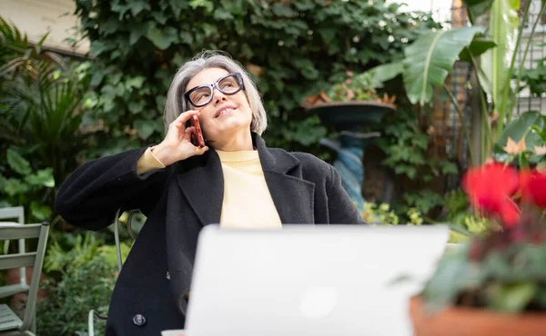 Dışarıda dizüstü bilgisayar üzerinde çalışırken akıllı telefonla iletişim kuran olgun bir kadın.