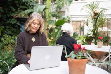 Yeşil bir bahçedeki masada dizüstü bilgisayar kullanan olgun bir kadın.