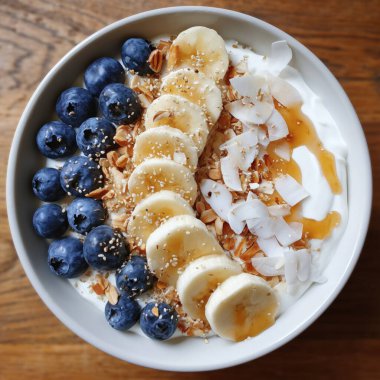 Yoğurt kasesi kahvaltıda yabanmersini, muz dilimleri, hindistan cevizi gevreği ve bal yağmuru ile hazırlanıyor.
