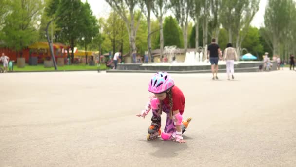 Una piccola pattinatrice si alza dalle ginocchia dopo una caduta, in un vicolo in un parco con una fontana. — Video Stock