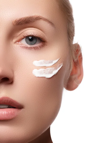 El rostro hermoso de la mujer joven con la crema cosmética en la mejilla. Concepto de cuidado de la piel. Retrato de primer plano aislado en blanco. Close-up joven hermosa cara de niña aplicando crema hidratante . —  Fotos de Stock