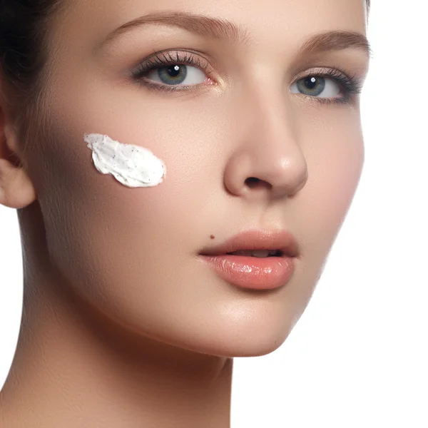 Bellissimo viso di giovane donna con crema cosmetica su una guancia. Il concetto di cura della pelle. Ritratto da primo piano isolato su bianco. Primo piano giovane bel viso di ragazza che applica crema idratante — Foto Stock