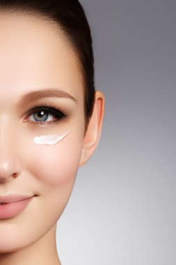 Beautiful face of young woman with cosmetic cream on a cheek. Skin care concept. Closeup portrait on beige background. Close-up young beautiful face of girl applying moisturize cream