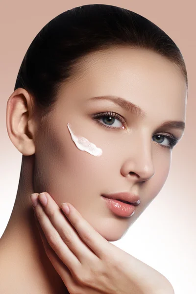 Beautiful face of young woman with cosmetic cream on a cheek. Skin care concept. Closeup portrait on beige background. Close-up young beautiful face of girl applying moisturize cream — Stock fotografie