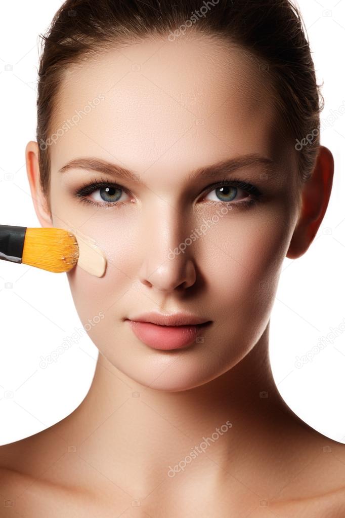 Beautiful face of young woman with cosmetic foundation on a skin. Beauty treatment concept. Makeup artist applying liquid tonal foundation on the face of the woman. Closeup photo of cheek