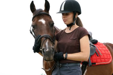 Kasklı ve binicilik ekipmanlı bir kız çarpık atıyla iletişim kuruyor. At kahverengi deri bir dizgin, eyer ve kırmızı bir eyer bezi giyiyor.