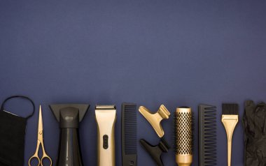 Mavi arka planda siyah ve altın rengi kuaför aletleri. Kuaför salonu aksesuarları, makas, toka, tarak, eldiven, saç kurutma makinesi ve yüz maskesi. Saç için düz eşyalar.