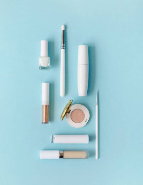 Mavi arka planda beyaz makyaj malzemeleri. Masanın üzerinde gölgeler, rimel, fırçalar, ruj, üst manzara var..
