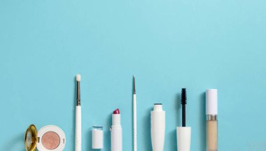 Mavi arka planda fotokopi alanı olan beyaz makyaj malzemeleri. Masanın üzerinde gölgeler, rimel, fırçalar, ruj, üst manzara var.