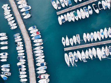 Ahşap rıhtımlar boyunca demirlemiş tekne ve yatlarla dolu bir marinanın havadan görüntüsü. Turkuaz su ve geometrik düzenleme temiz, modern ve görsel olarak çekici bir bileşim oluşturur.