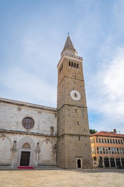 Slovenya 'nın Koper kentindeki tarihi Varsayım Katedrali ve etkileyici taş çan kulesinin ayrıntılı bir görüntüsü. Ortaçağ mimarisi, süslemeler ve saat kulesi parlak mavi gökyüzünün altında duruyor.