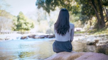 Ormandaki sakin bir nehrin kıyısında, kayaların üzerinde oturan kadının arka görüntüsü, yumuşak ışık, açık hava rahatlığı ve tefekkür ile huzurlu doğa sahnesi..