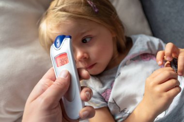 Anne, evde kızılötesi termometresi olan çocuğu için ateşini ölçüyor. Annesi dijital kızılötesi termometreyi tutuyor..