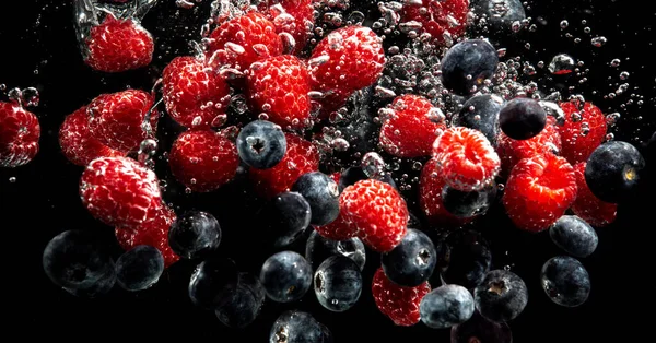 Blueberries and raspberries splashing into water with air bubbles and sinking on black — Stock Photo, Image