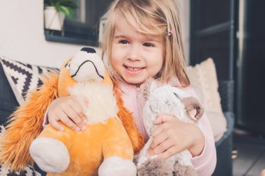 Küçük kız gülümsüyor ve sarılıyor iki peluş oyuncak köpek ve kedi.