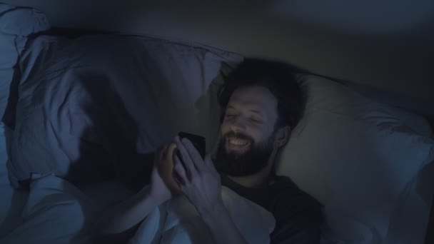 Гаджет ніч розслаблений чоловік прокручує телефон в ліжку — стокове відео