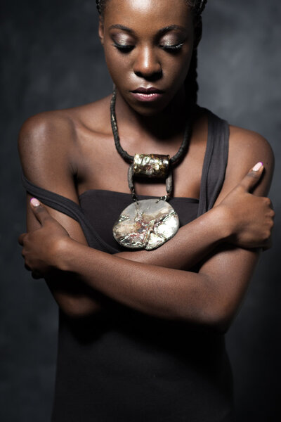 The sad black woman with an ethnic ceramic brilliant necklace embraces herself for shoulders and looks down. Studio photo against a dark background