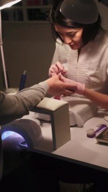 Dikey video. El manikürü. Tırnaklar önemser. Manikürcü kadın müşterileri kaplıyor. Manikür lambasında kurutan üst kat sıvısı..