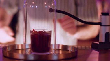 Kokteyl barı. Barmen ekipmanları. Büyük cam saatin altında dumanı tüten bir silahla kırmızı alkol içeceği hazırlıyorum. Bar tezgahında şık bir sunum.