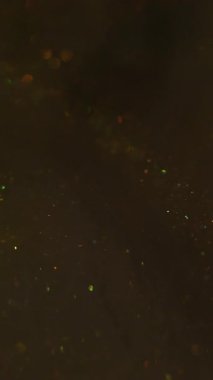 Dikey video. Hipnoz sıvısı. Sanat pırıltısı. Parıldayan karışım parçacıkları bokeh kıvılcımlarında harmanlıyor. Renkli altın metalik boya akışı soyut odaklanmış siyah arkaplan.