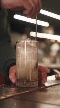 Dikey video. Kokteyl yapımı. Buzlu içecek. Barmen uzun bardakta uzun metal kaşıkla köpüklü içecek karıştırıyor. Profesyonel bar aletleri.