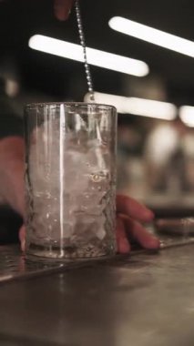 Dikey video. Soğuk içecek hazırlanıyor. Buz karıştırıyorum. Barmen el tekniği, uzun şeffaf bardakta donmuş suyu karıştırıyor. Serinletici kokteyller için soğutma.