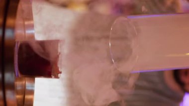 Dikey video. Barmen gösterisi. İçki sunumu. Erkek barmen dumanlı kırmızı alkollü kokteylden cam kaput kaldırıyor. Güzel bir buhar barı tekniği..