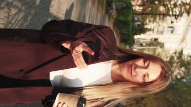 Dikey video. Konuşan kadın. Modern teknoloji. Kahveli, mutlu, şık bir iş kadını dışarıda telefonla konuşuyor..