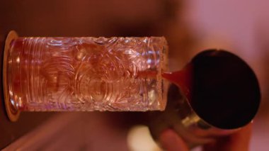 Dikey video. Kokteyl dolduruyorum. Soğuk aperatif. Barmen el metal karıştırıcıdan desenli cama kırmızı sıvı ekliyor. Barda buz küpü içeceği yapıyorum..