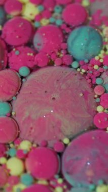 Dikey video. Akışı kes. Renk sıvısı. Odaklanmamış pembe, sarı, parlak mürekkep damlaları soyut arka planda süzülen parlak pigmentler..