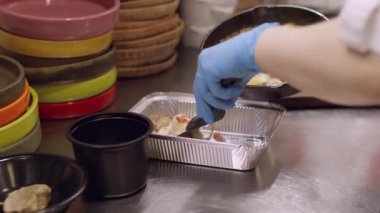 Yemek dağıtım süreci. Paket yemek hazırlıyorum. Şef eldivenlerini giyip, spatulayla pişirilmiş sosu folyo kabına koyuyor. Depo için paketleme kabı.