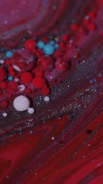 Dikey video. Akışı kes. Renk sıvısı. Odaklanmamış kırmızı mor parlak mürekkep damlacıkları soyut arkaplanda parıldayan parlak pigmentler..