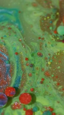 Dikey video. Akışı kes. Renk sıvısı. Odaklanmamış yeşil mavi parlak mürekkep köpükleri soyut arkaplanda parlayan turuncu pigmentler..