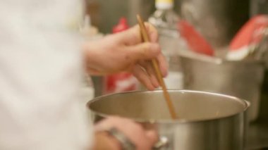 Aşçılık işi. Karışık yemek. Beyaz üniformalı aşçı sıcak yemeği metal tencerede ahşap spatulayla karıştırıyor. Tencerede buharlı yemek pişirmek..