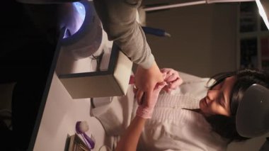 Dikey video. El manikürü. Tırnaklar önemser. Manikürcü kadın müşterileri kaplıyor. Manikür lambasında kurutan üst kat sıvısı..