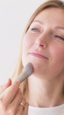 Dikey video. Günlük makyaj malzemesi. Dekoratif kozmetik. Kozmetik fırça kullanan tatmin olmuş kadın beyaz tenli kadın yüzüne yüz sürüyor..