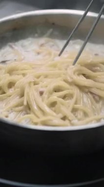 Dikey video. Spagetti pişiriyorum. Erişte yemeği. Şef sıcak vejetaryen tavasını fırında metal cımbızla karıştırıyor. Beyaz soslu lezzetli makarna..