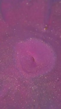 Dikey video. Köpüklü serpinti. Mürekkep damlası. Soyut akrilik arkaplan üzerinde pembemsi mor, altın renkli, parlak sıvı halkalar..