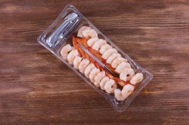 Kahverengi ahşap bir masada plastik bir kabın içinde soyulmuş ve pişmiş karides.