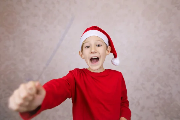穿着红色夹克 头戴圣诞帽 背景轻盈 快乐的男孩 免版税图库图片